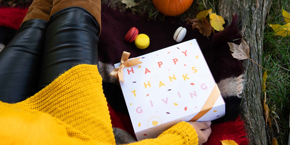 A woman in a yellow sweater is sitting on a floor full of leaves and pumpkins. On her hand is a French macaron box with a Thanksgiving sleeve.
