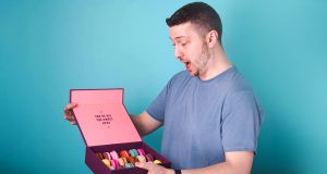 A man with a surprised expression is holding a box full of assorted French macarons in his hands.