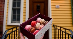 A box full of 9 French macarons in front of a yellow house.