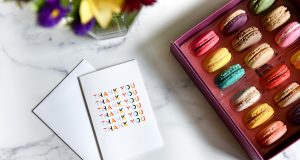 A box full of French macarons has a thank you greeting card and some flowers to the left.