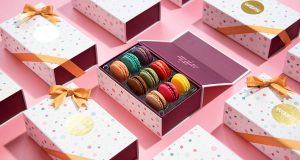 A box full of French macarons is surrounded by French macaron boxes with golden ribbons.