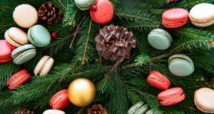 Assorted French macarons on top of pine tree branches surrounded by holiday ornaments. 