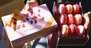 A pair of hands hold a box of 9 macarons and a Valentines Day box sleeve
