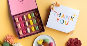 A box full of French macarons has a macaron box with a thank you sleeve to the right. Below is a plate full of macarons and some flowers