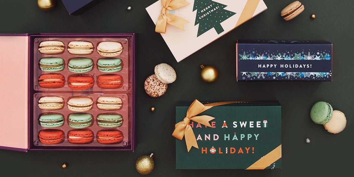 A box full of French macarons is surrounded by macaron boxes with holiday sleeves, holiday ornaments, and macarons.