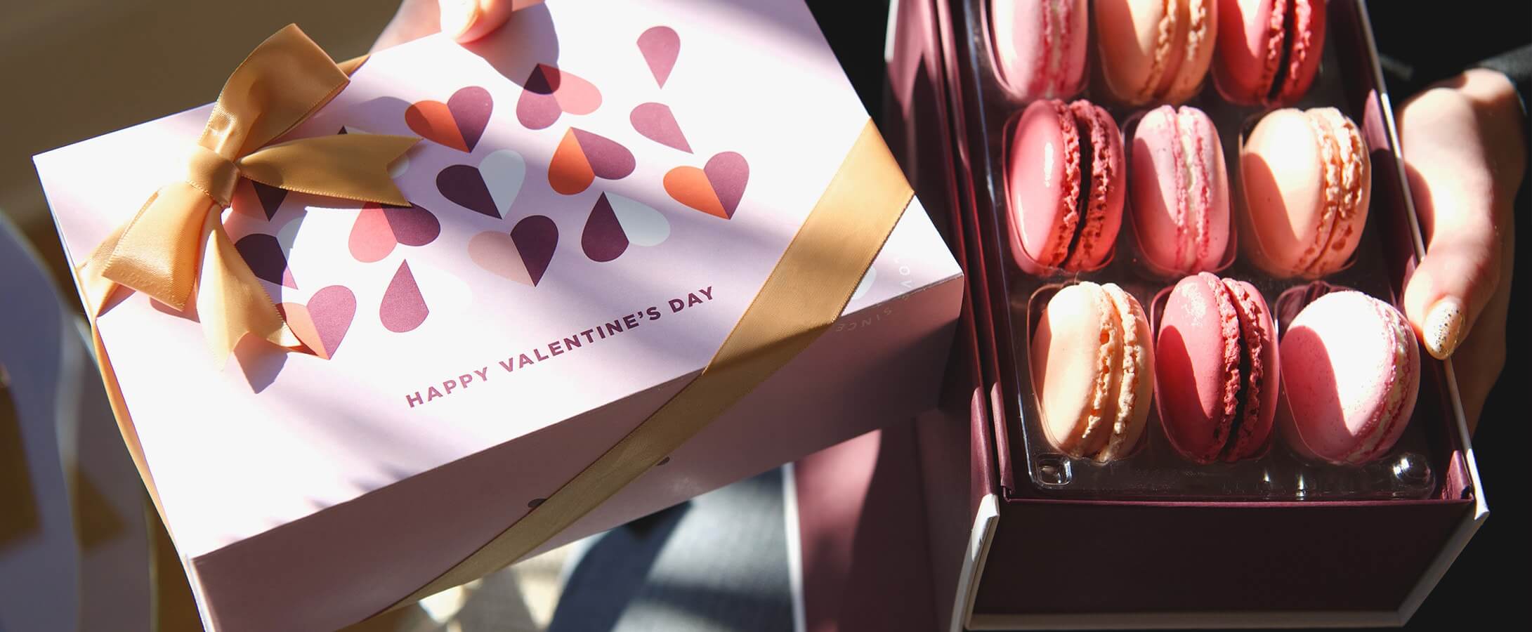 A box full of macarons and a macaron box with a Valentine’s Day sleeve are being held by female hands.