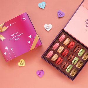 A box full of eighteen assorted macarons has a macaron box with a Valentine's Day sleeve to the left. Surrounding them are some candy hearts.