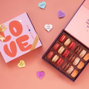 A box full of eighteen assorted macarons has a macaron box with a Valentine's Day sleeve to the left. Surrounding them are some candy hearts.