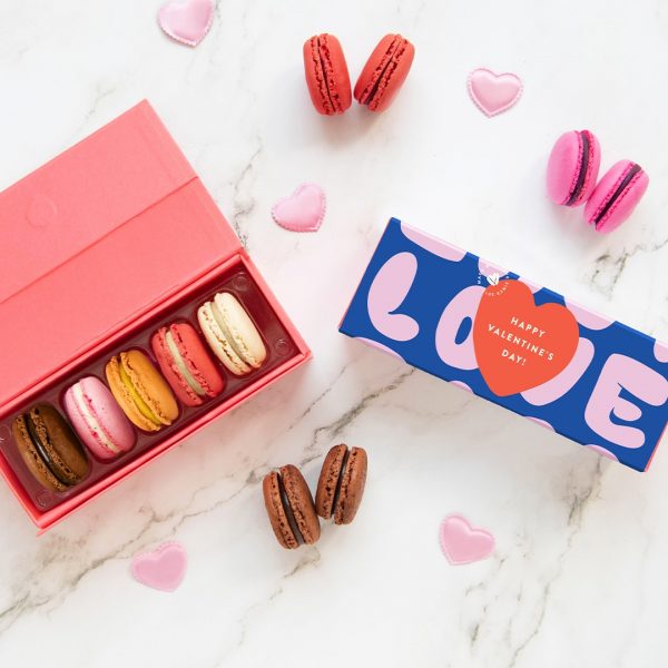 A box with five French macarons has a macaron box with a Valentine's Day sleeve. Surrounding them are some assorted macarons and hearts.