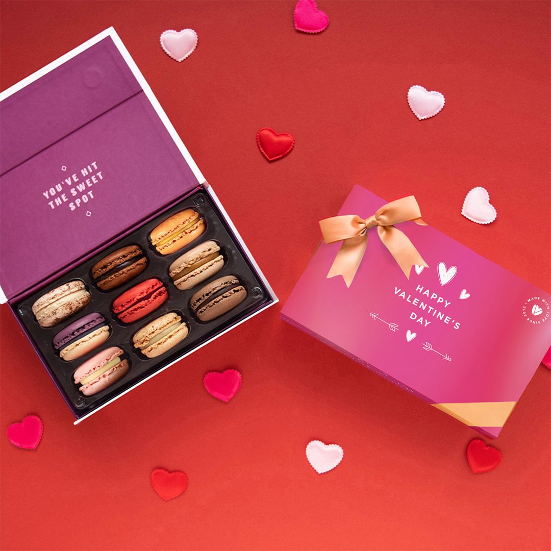 Build Your Own Full Heart Box - 1080x10802026 Close-up of an open box of macarons with "You've Hit the Sweet Spot" message, next to a "Valentine's Day" themed box with a bow, on a red surface with scattered heart confetti.