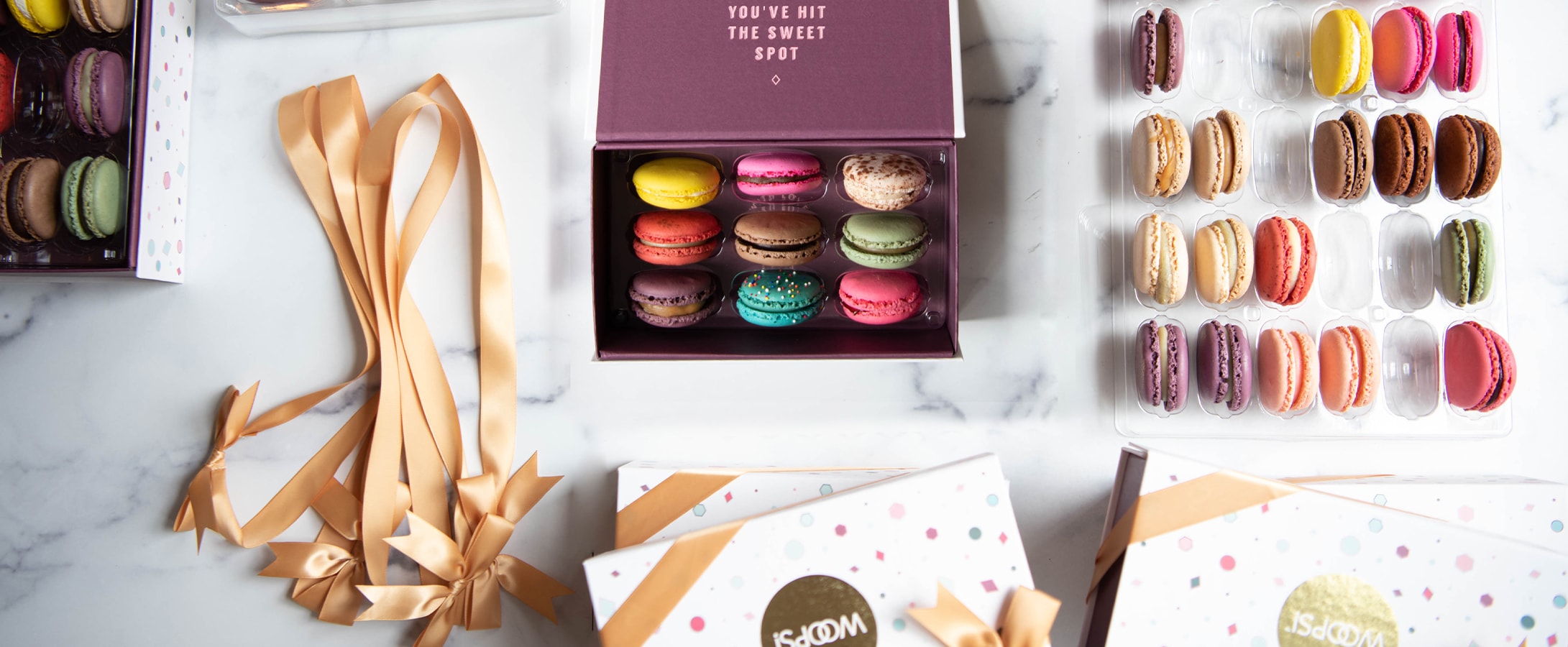 A tray and a box full of French macarons are surrounded by macaron boxes and golden ribbons.