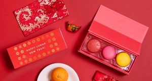 A box with three French macarons has a macaron box with a Lunar New Year sleeve to the left. Surrounding them are some tangerines, red envelopes, and a golden Chinese ornament. 