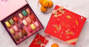 A box full of 18 assorted macarons has a macaron box with a Lunar New Year sleeve to the right. Surrounding them are some tangerines on a plate, a red envelope, and two golden Chinese ornaments.