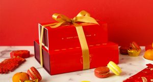 A macaron stack with Lunar New Year sleeves is surrounded by assorted macarons, red envelopes, and tangerines. 