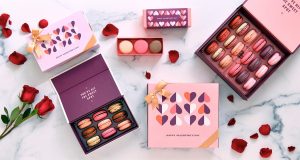 Three boxes full of macarons are surrounded by macaron boxes with Valentine’s Day sleeves, red rose petals, and two red roses