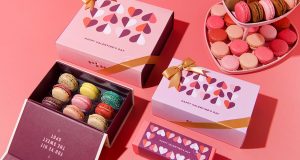 A box and a tiered tray full of French macarons have macaron boxes with Valentine’s Day sleeves between them. 
