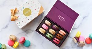 A box full of French macarons has a macaron box with a golden ribbon to the left. At the bottom are some assorted macarons.