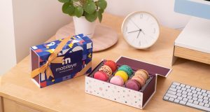 A box full of French macarons has a macaron box to the left. Behind is a clock, a computer, and a plant. 