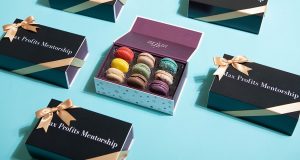 A box full of French macarons is surrounded by macaron boxes with black sleeves.
