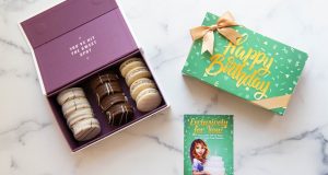 A box full of assorted alfajores is surrounded by macaron boxes with green custom sleeves.