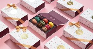 A box full of assorted macarons is surrounded by macaron boxes with golden ribbons. 
