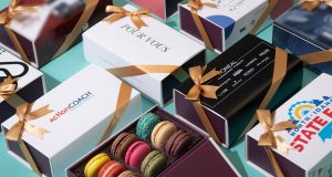 A box full of assorted French macarons is surrounded by Woops! macaron boxes with different customized sleeves.