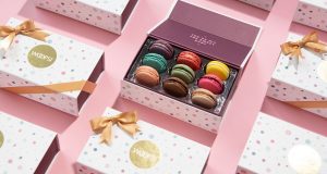 A box full of assorted French macarons is surrounded by Woops! macaron boxes with golden ribbons.