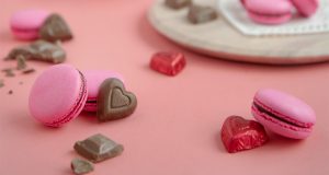  Several pink Milk Chocolate macarons are on plates and lying around. Surrounding them are some heart-shaped chocolates.