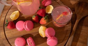 Numerous pink Strawberry Lemonade macarons are surrounded by strawberries and a glass and jar filled with strawberry lemonade.