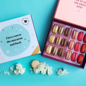 A blue background features a Woops! box of 18 French macarons with assorted flavors, to its left is a white and blue sleeve with a golden ribbon and some white flowers at the bottom.