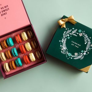 A green background features a Woops! box of 18 French macarons with assorted flavors and a dark green sleeve with a golden ribbon and an ornament figure at the center.