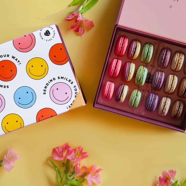 A box full of 18 assorted French macarons has a macaron box with a Just Because sleeve to the left. Surrounding them are some flowers