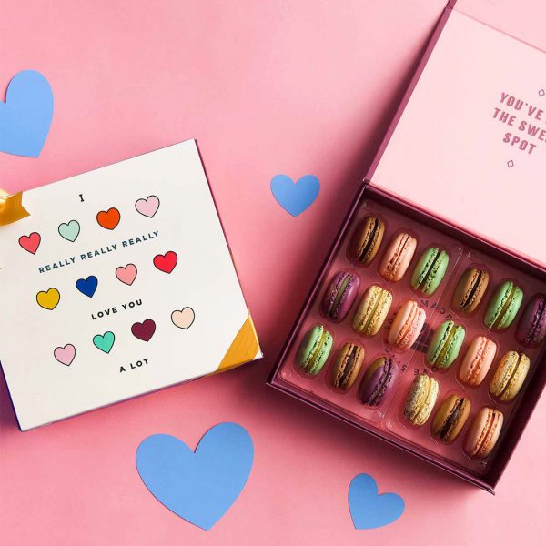 A box full of 18 assorted French macarons has a macaron box with an I Love You sleeve next to it. Surrounding them are some blue paper hearts.