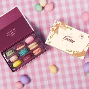 Open box of colorful French macarons next to a cream “Happy Easter” gift box with ribbon, surrounded by pastel Easter eggs on a pink gingham background.