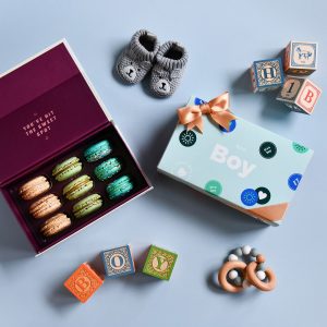 A box full of 9 assorted French macarons has a macaron box with a Baby Shower sleeve to the right. Surrounding them are some baby boy toys.