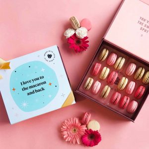 A box full of 18 assorted French macarons has a macaron box with an I Love You sleeve to the left. At the top and bottom are some macarons and flowers.