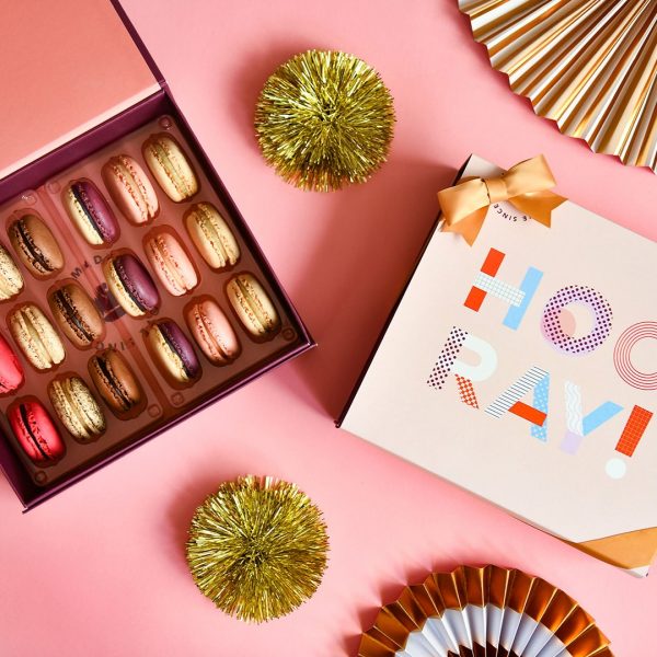 A box full of 18 assorted French macrons has a macaron box with a Congratulations sleeve to the right. Surrounding them are some golden party decorations.