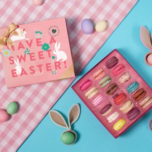 A box full of 18 assorted French macarons has a macaron box with a blue Easter sleeve to the left. Surrounding them are some colored eggs and bunny ears.