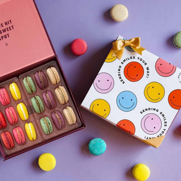 A box full of 18 assorted French macarons has a macaron box with a smiley faces sleeve to the right. Surrounding them are some assorted macarons.