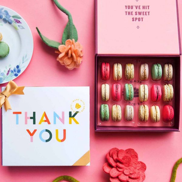 A box full of 18 assorted French macarons has a macaron box with a Thank You sleeve next to it. At the top is a plate with macarons and a flower and at the bottom a big pink flower.