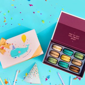 A box full of nine assorted French macarons has a macaron box with a colorful Happy Birthday sleeve to the left. Surrounding them are some party decorations and tiny candles.
