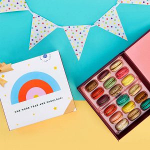 A blue and yellow background features a Woops! box of 18 French macarons with assorted flavors to the right and a macaron box with a red sleeve to the left. At the top is a party decoration.