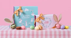 Two Woops! French macaron boxes with Easter sleeves are surrounded by assorted macarons, colorful eggs, and bunny ears