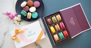 A Woops! box full of assorted French macarons has a macaron box with an Eid sleeve and a plate full of assorted macarons to the left