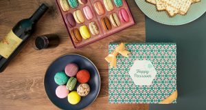 A Woops! box full of 18 assorted French macarons has a plate full of macarons and a macaron box with a Passover sleeve below. It also has a plate with matzos to the right and a bottle of wine to the left. 