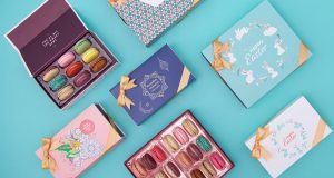 Two boxes full of assorted macarons are surrounded by French macaron boxes with colorful Ramadan, Easter, Blossoming Colors, and Passover sleeves. 