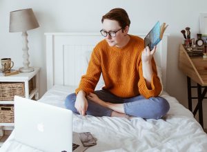 A smiling woman in an orange sweater is sitting in her bed looking at her computer screen. Behind her is a white wall with three hats, a bedside table with a lamp on it, and a desk with some stuff lying around. 