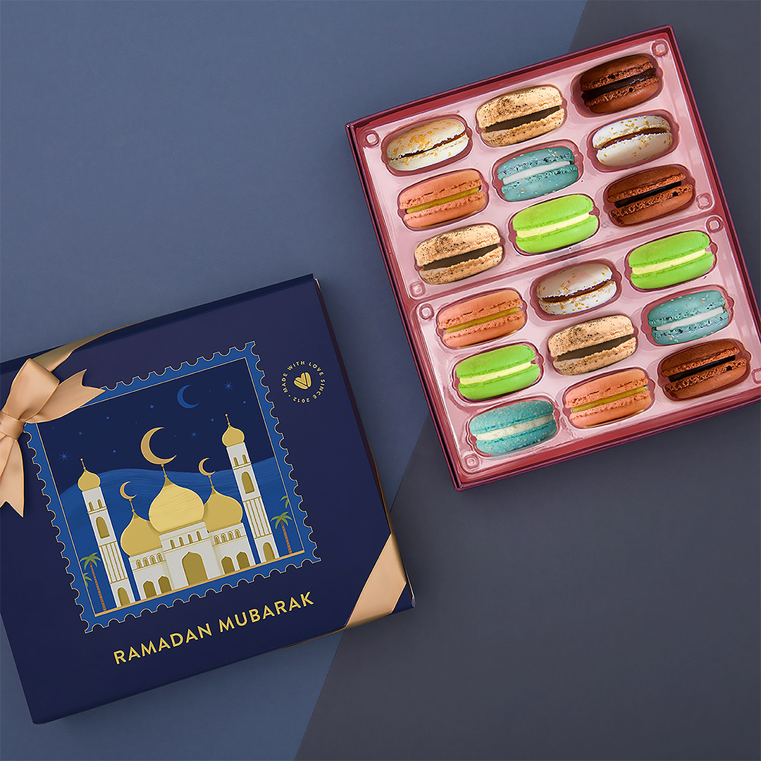 New Crescent Moon - 1080x10802026 A box of 18 French macarons with assorted flavors has a box of French macarons with a dark blue Ramadan sleeve to its left.