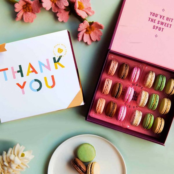A box full of 18 assorted French macarons has a macaron box with a Thank You sleeve to the left. At the bottom is a plate with macarons and at the top some pink flowers.