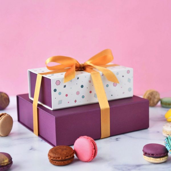 A macaron box of 9 on top of a box of 18 with a golden ribbon holding them together, and some assorted French macarons lying around.
